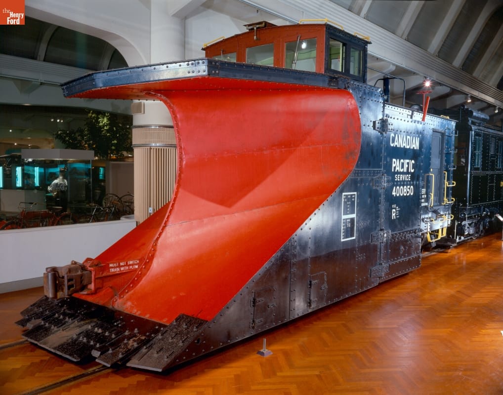 Canadian Pacific Snowplow, 1923 Black railcar with scooped-out front painted red, sitting on tracks in a large room
