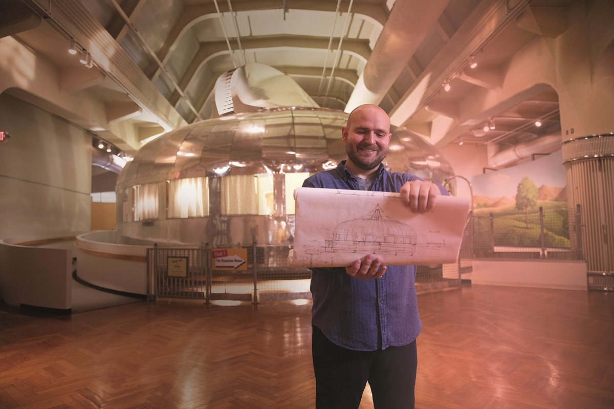 Ten-year The Henry Ford member Blake Almstead Man stands in large building with round silver metal structure behind him, holding a drawing of the same structure