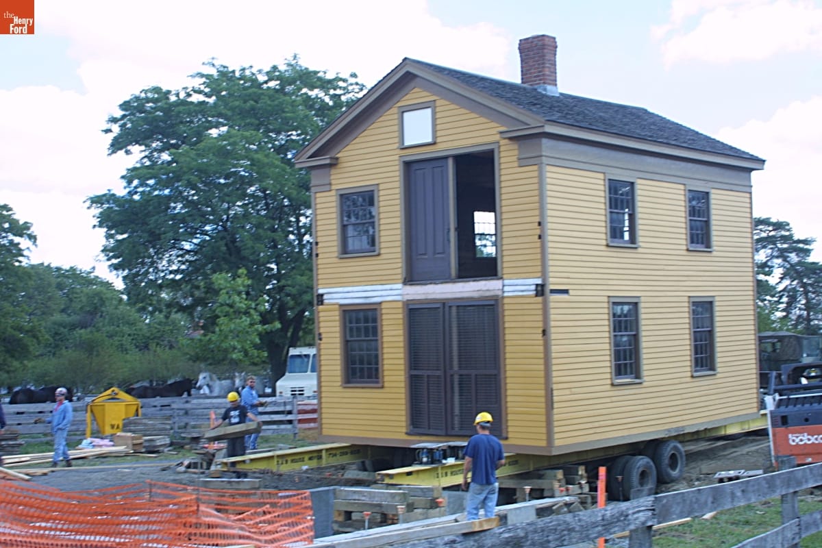 Two-story yellow wooden building on rails and wheels on a construction site with people in hard hats nearby 