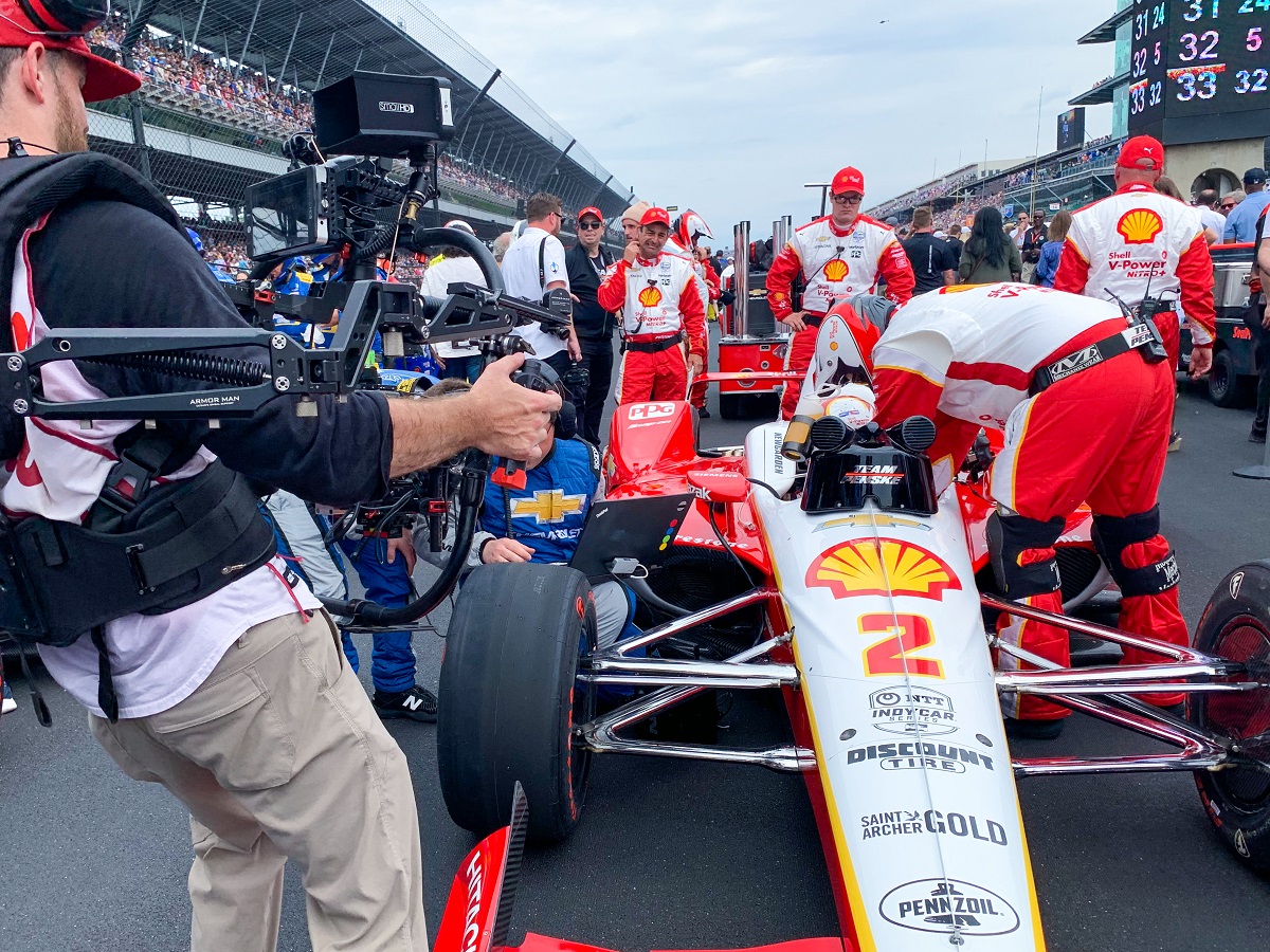 Man holding camera films long, low race car with pit crew in uniforms standing nearby and crowd in grandstands to both sides