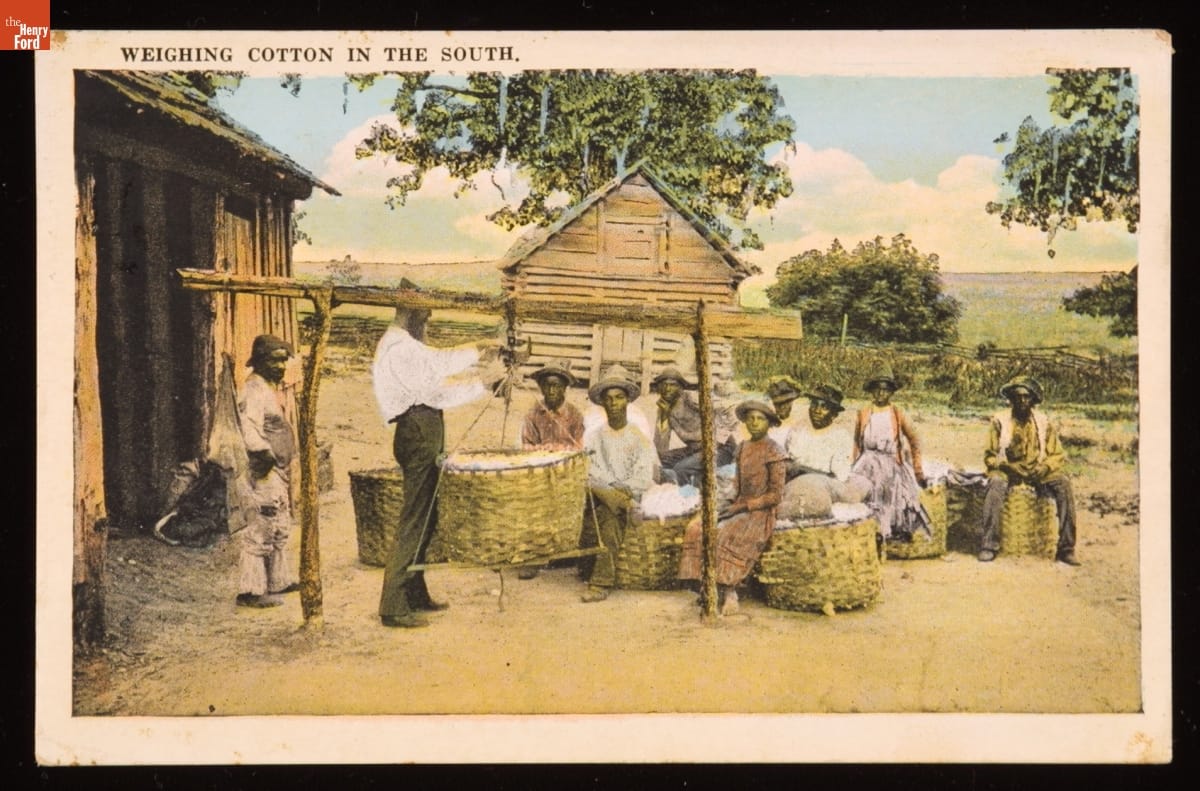 Man hooks large basket up to a wooden framework in a clearing among buildings and trees as other people with baskets sit nearby
