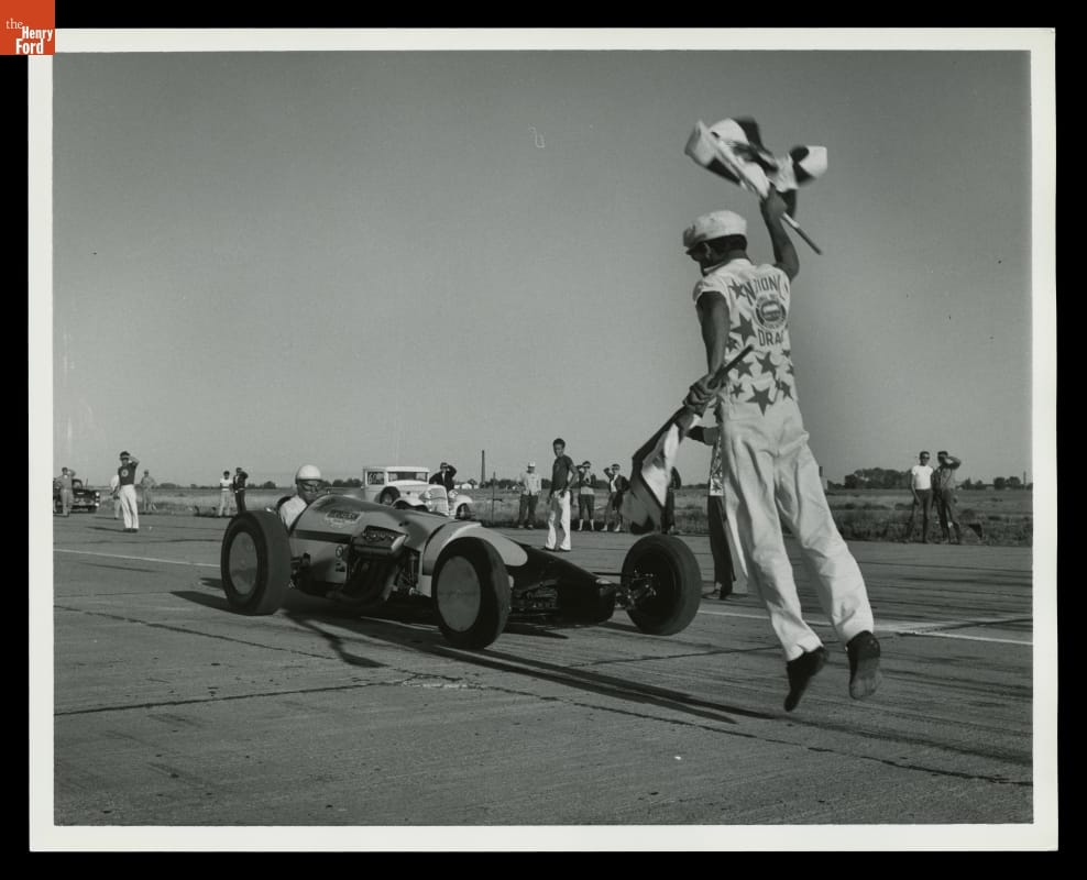 Official Start of First NHRA Drag Racing Meet, Great Bend, Kansas, 1955 Person holding a checkered flag in each hand in mid-jump in front of a race car on a track