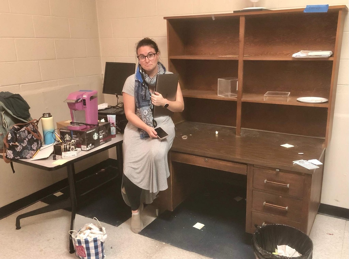 Jillian in the old Photo Studio Frowning woman sits on edge of desk with hutch next to table filled with bags and boxes