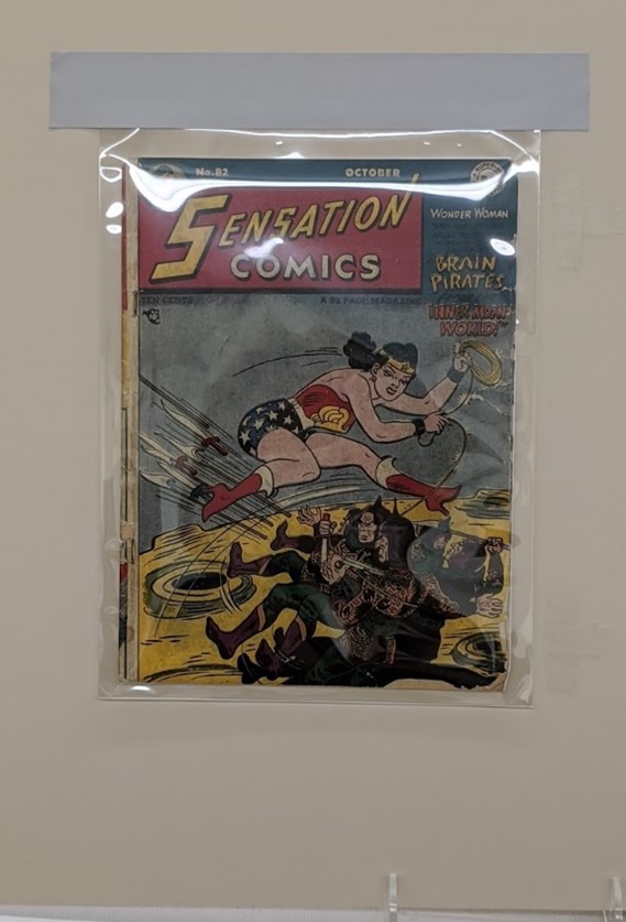 Comic book featuring Wonder Woman lassoing two other figures, under clear plastic in a mat