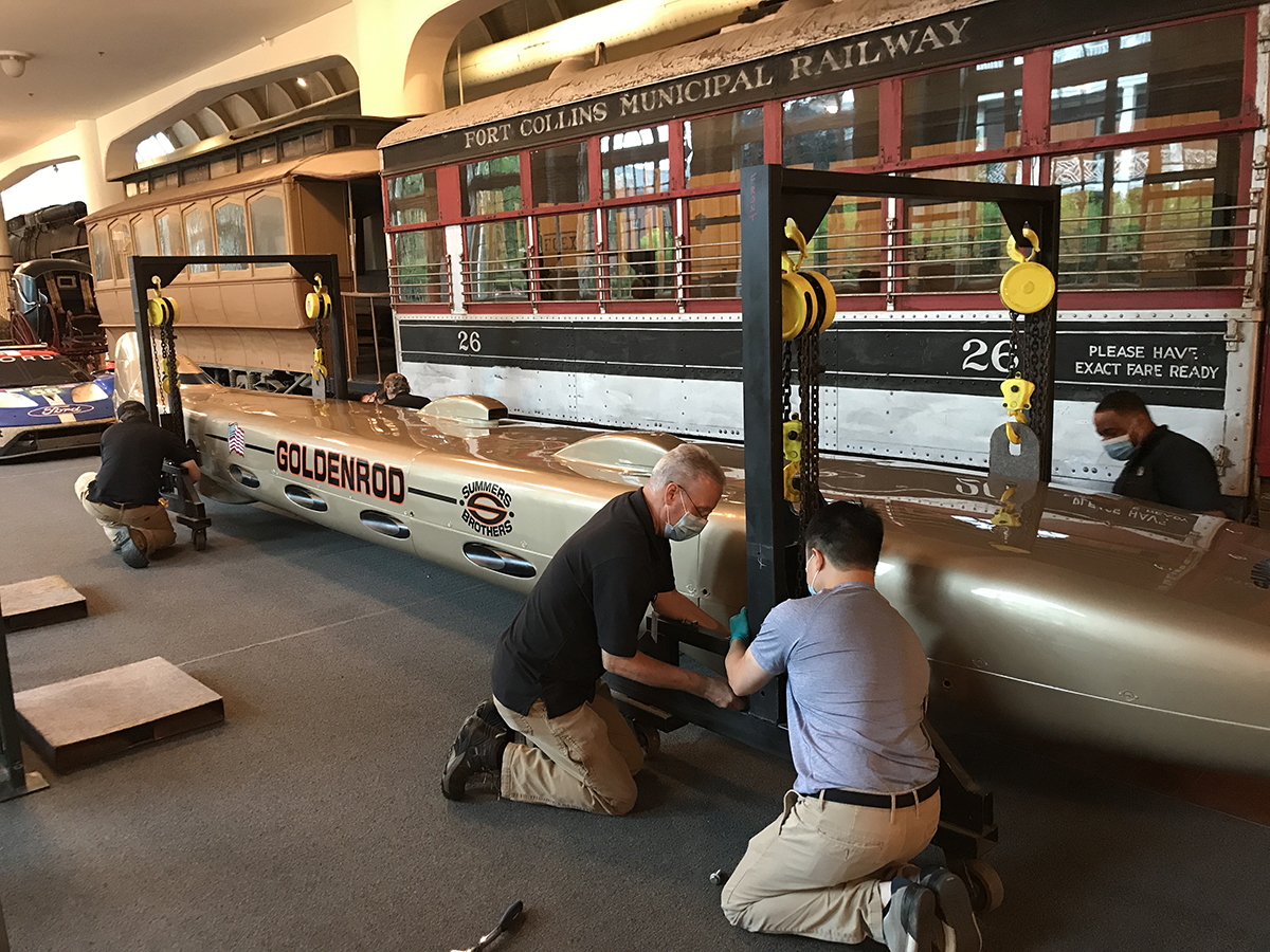 Conservation and Exhibits staff attach gantries to Goldenrod to enable movement. Men wearing face masks kneel around a long, low, torpedo-shaped golden car
