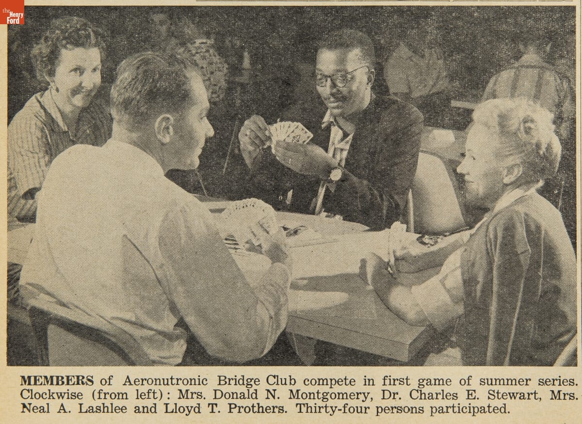 Black-and-white photo of four people sitting around a table playing cards; other similar tables of people visible in background