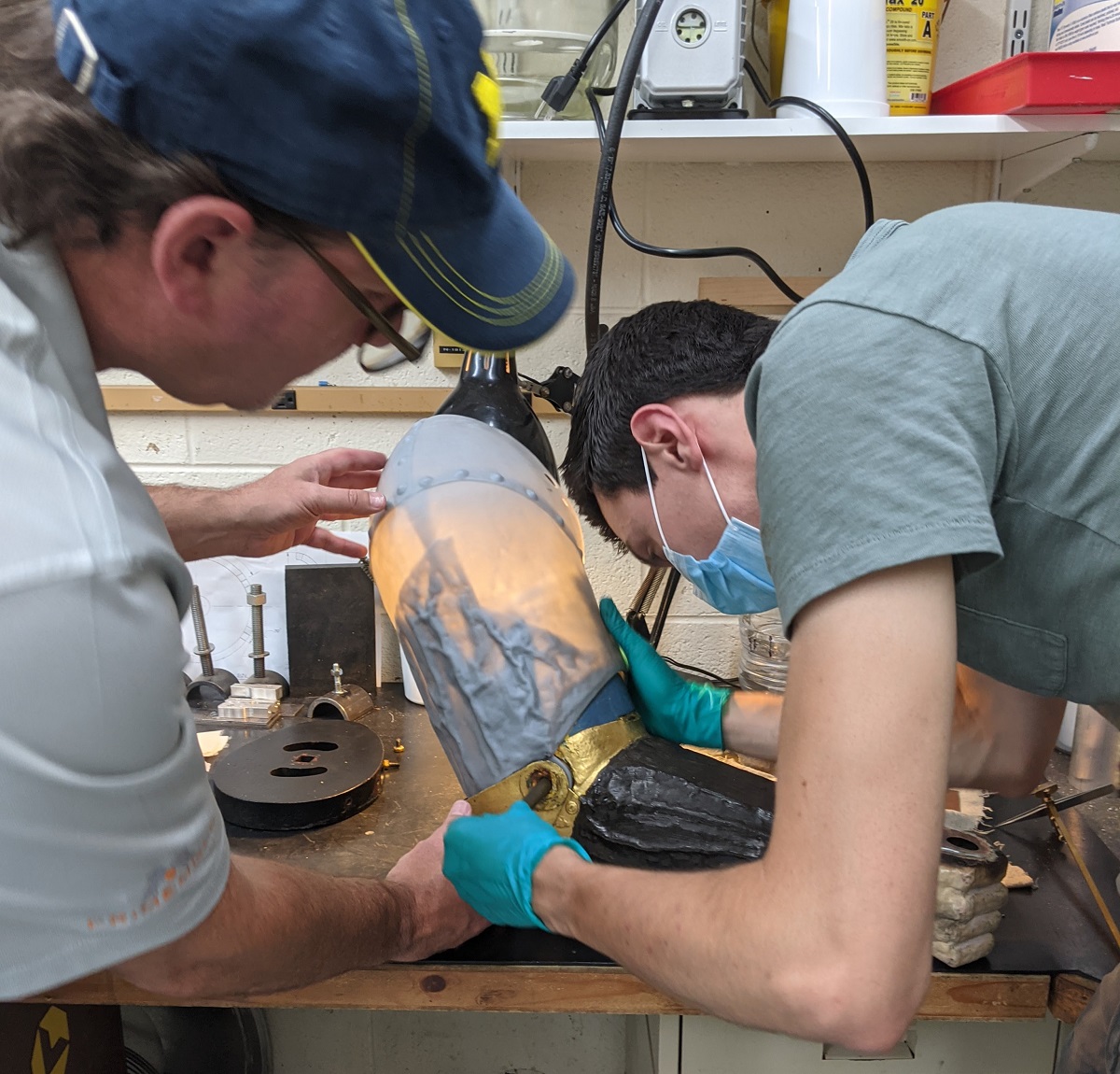 Two people handle a hollow statuary arm on a workbench