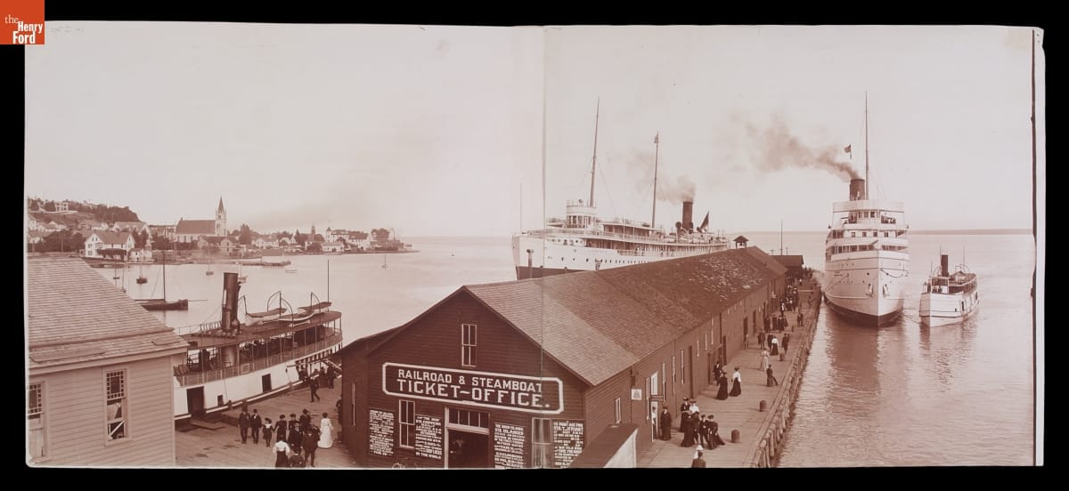 Black-and-white image of dock with long low building on it and several large ships in the water around it