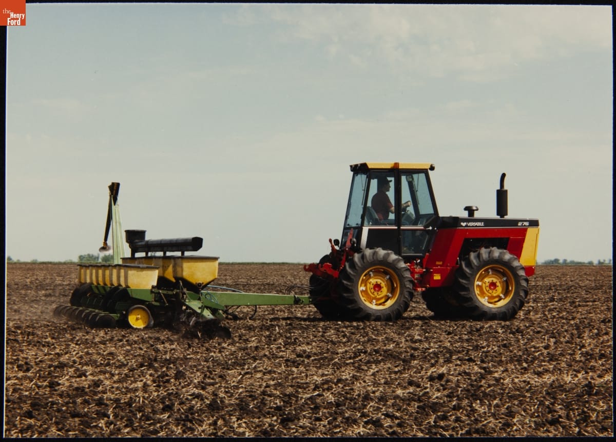 Small red tractor with enclosed cab pulling a planter through a dirt field