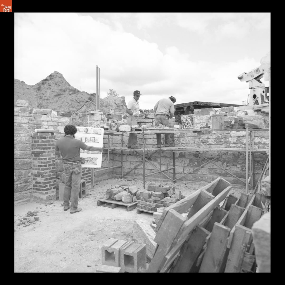 Using Photographs of the Original Walls to Reconstruct Firestone Farmhouse in Greenfield Village, August 1984 Two men construct a stone block wall on a construction site, while a third man looks at a large display board he is holding that contains photographs