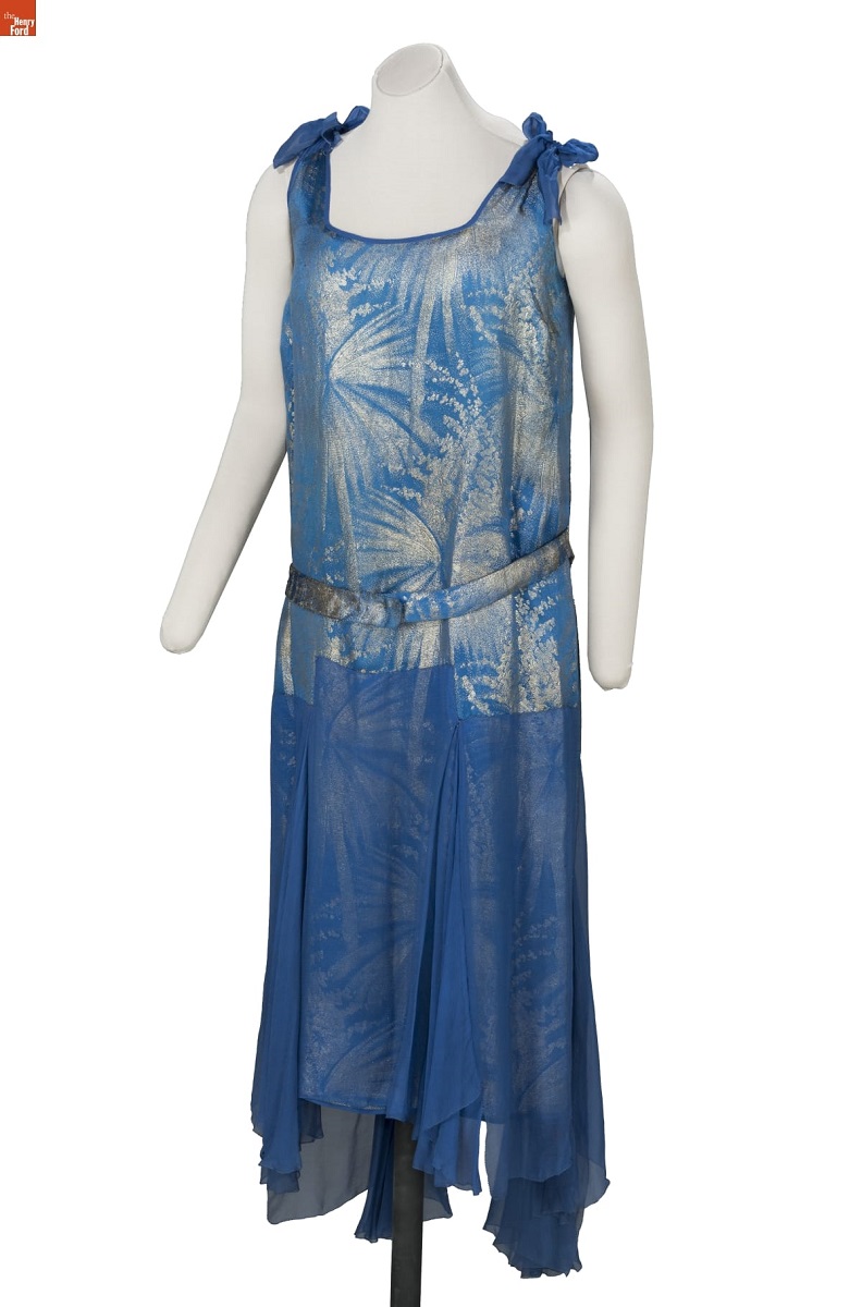 Mannequin wearing blue and silver sleeveless drop-waist shift dress with firework- or foliage-patterned top and solid blue handkerchief skirt