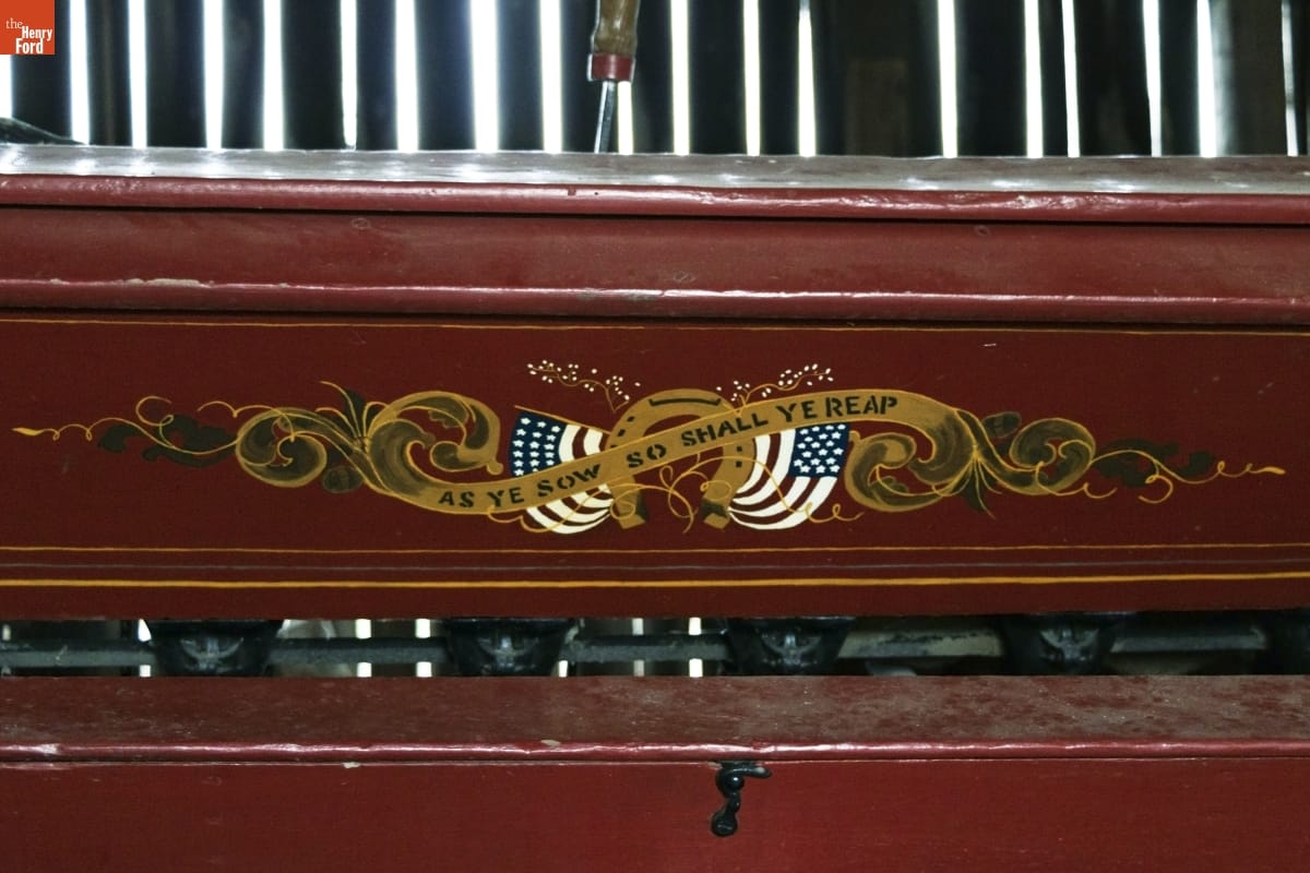 Brown or maroon wooden equipment with text "As Ye Sow So Shall Ye Reap" against decorative gold scrolling, two American flags, and a horseshoe 