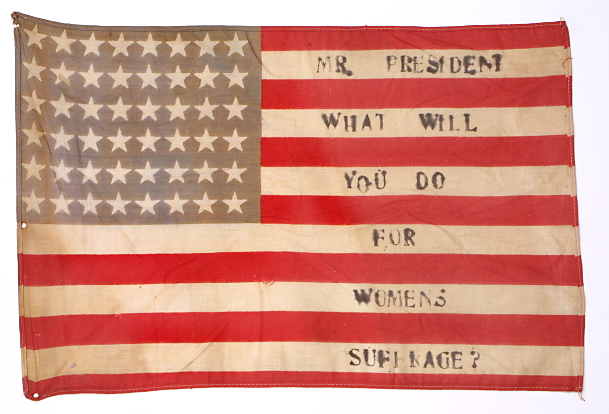 48-star American flag with red and white stripes and white stars on a blue field in upper left with text "Mr. President, What Will You Do for Women's Suffrage?" on white stripes