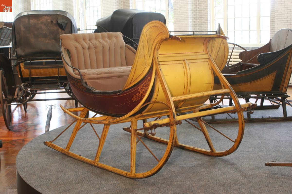 Yellow and maroon sleigh with beige upholstered seat, displayed in a room with other vehicles