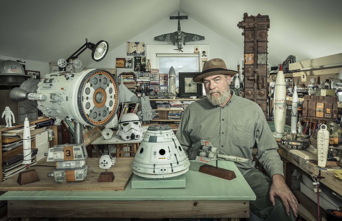 Man in fedora sits in a workshop crowded with models (mostly space-related), books, and other items