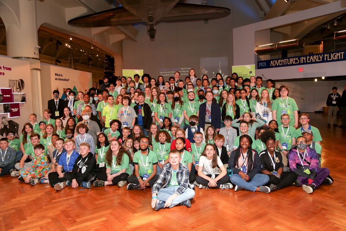 A group of children, many wearing green t-shirts and lanyards, pose on the floor of a museum