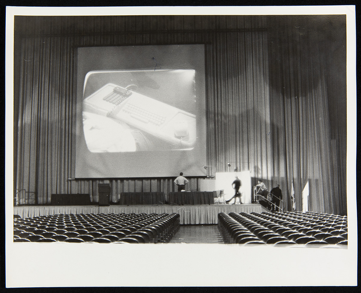 Fall Joint Computer Conference, San Francisco, California, December 1968 Empty auditorium with rows of seats and stage with curtains, a large screen with a piece of equipment projected on it, and several people on the stage
