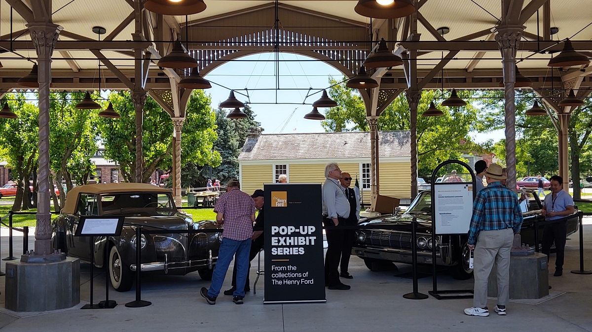Onlookers check out two cars behind stanchions with signage under a large open shed