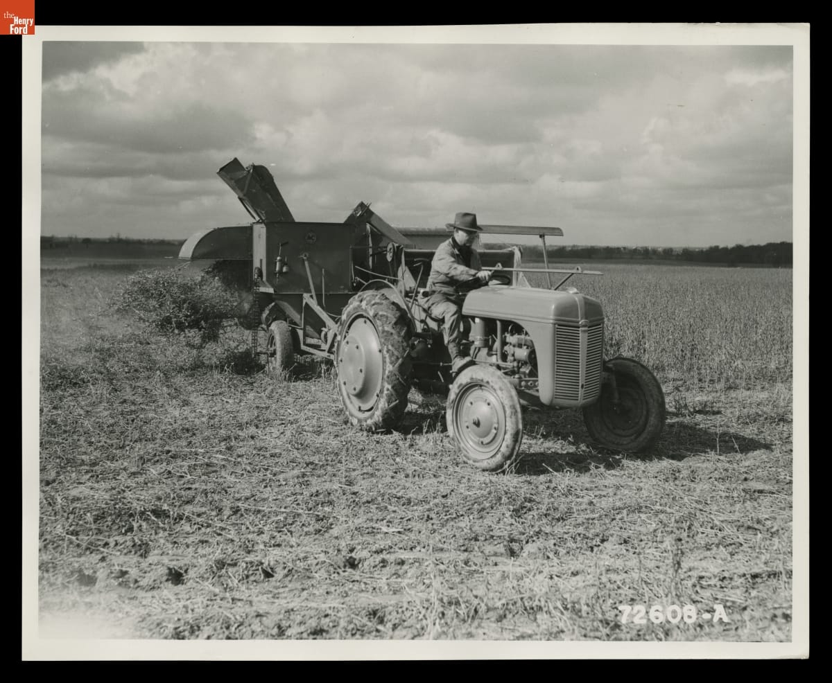 Black-and-white photo of a person riding a tractor through a field, pulling agricultural equipment containing crops behind it 