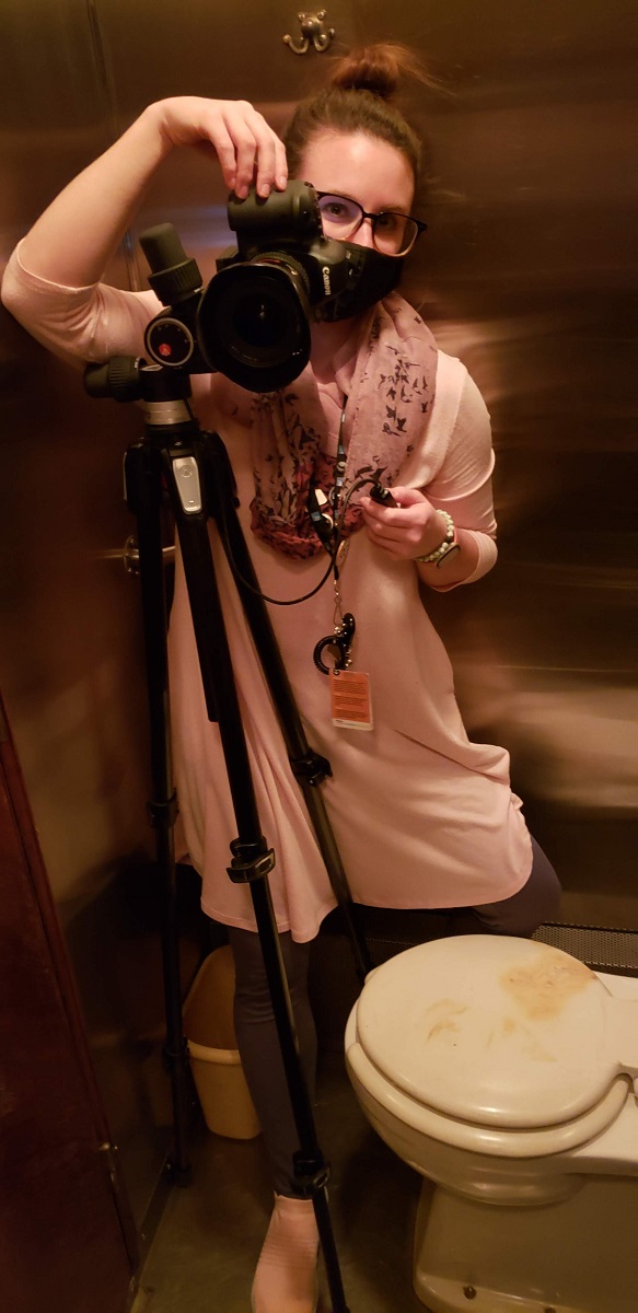 Digital Imaging Specialist Jillian Ferraiuolo in the bathroom of the Fair Lane railroad car Woman wearing mask behind camera on tripod in a stainless steel room containing a toilet
