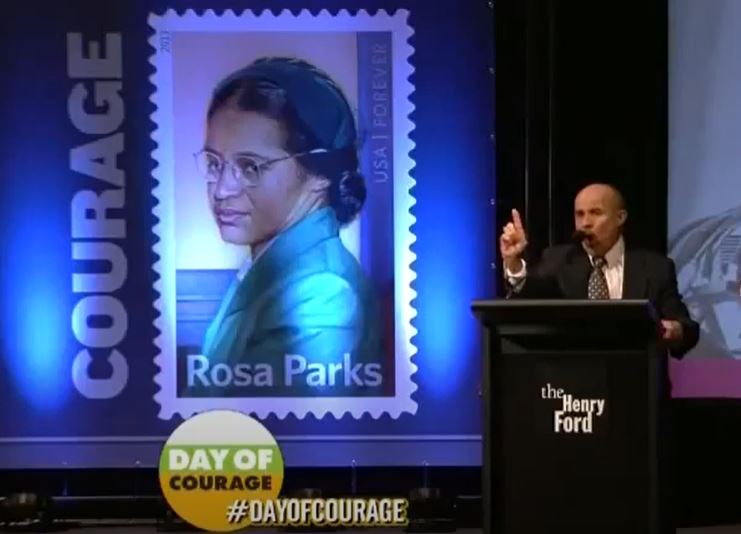Anthony reciting “I’ve Seen the Mountain Top” at National Day of Courage Man in suit stands at podium with hand pointed at the audience in front of backdrop of large Rosa Parks stamp