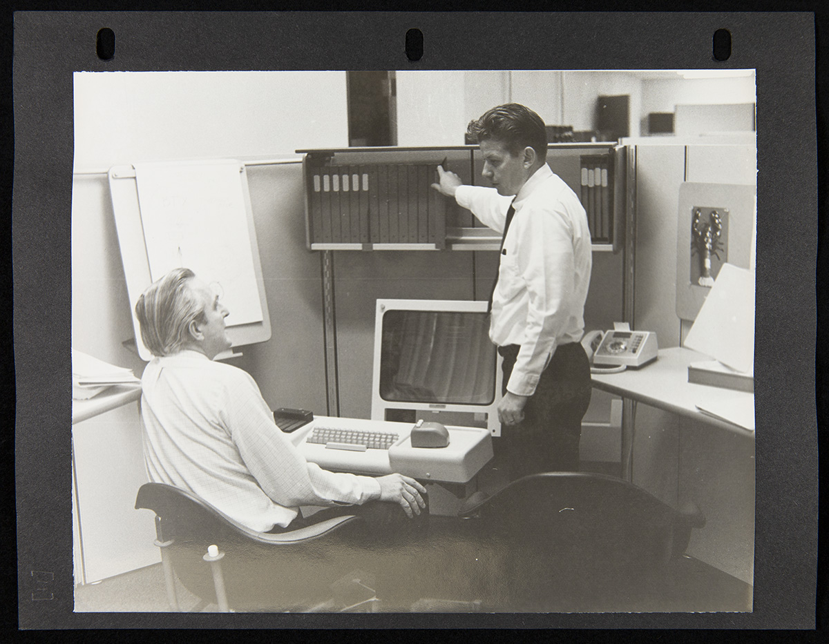 Page from Report on the SRI Fall Joint Computer Conference, December 27, 1968 Two men, one seated and one standing, among office furniture holding a computer, phone, and books or binders