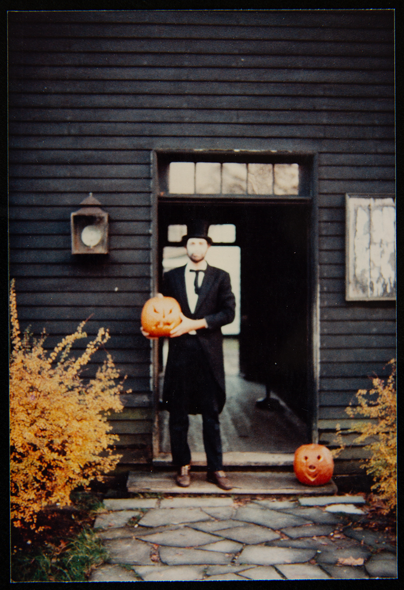 Man in long jacket and stovepipe hat in doorway holding a jack-o-lantern