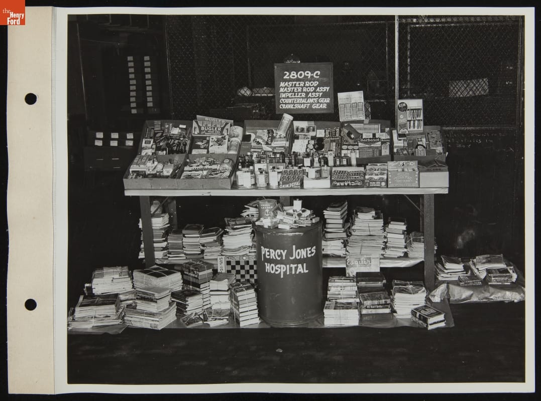 Table piled with small items, books and magazines underneath, along with bin labeled “Percy Jones Hospital”