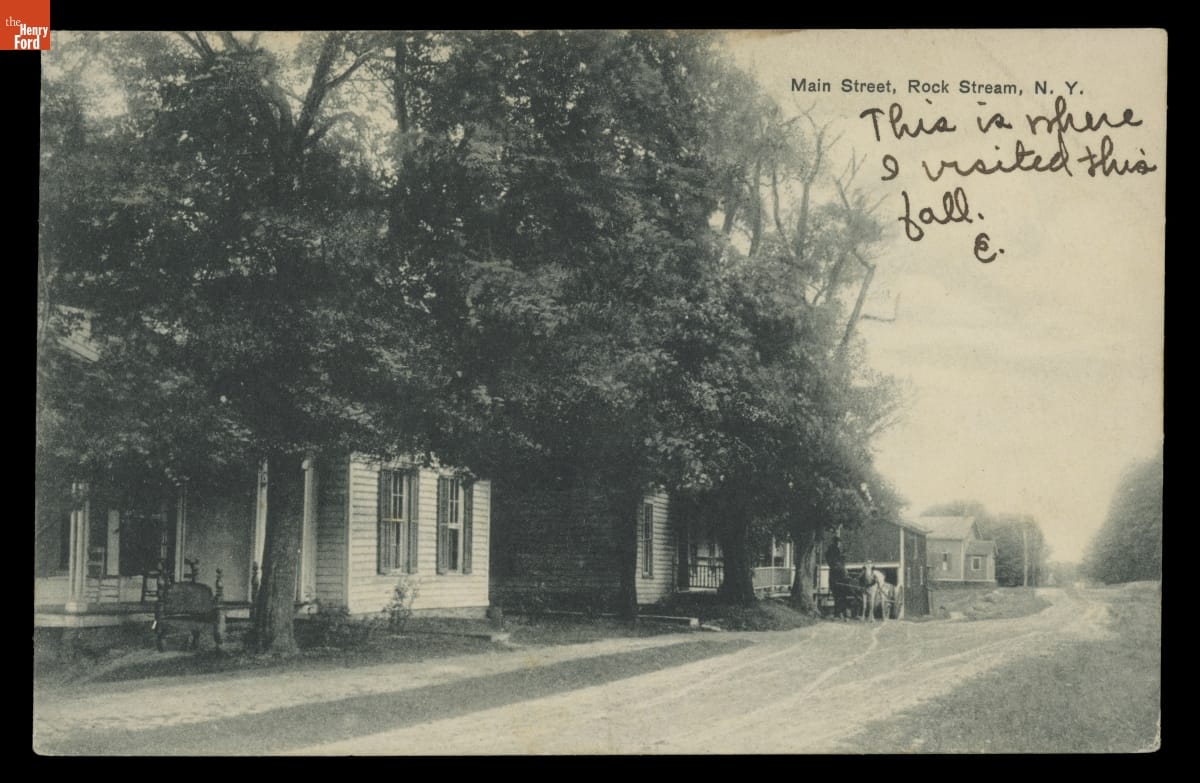 Black-and-white image of houses and trees along dirt road; printed and handwritten text