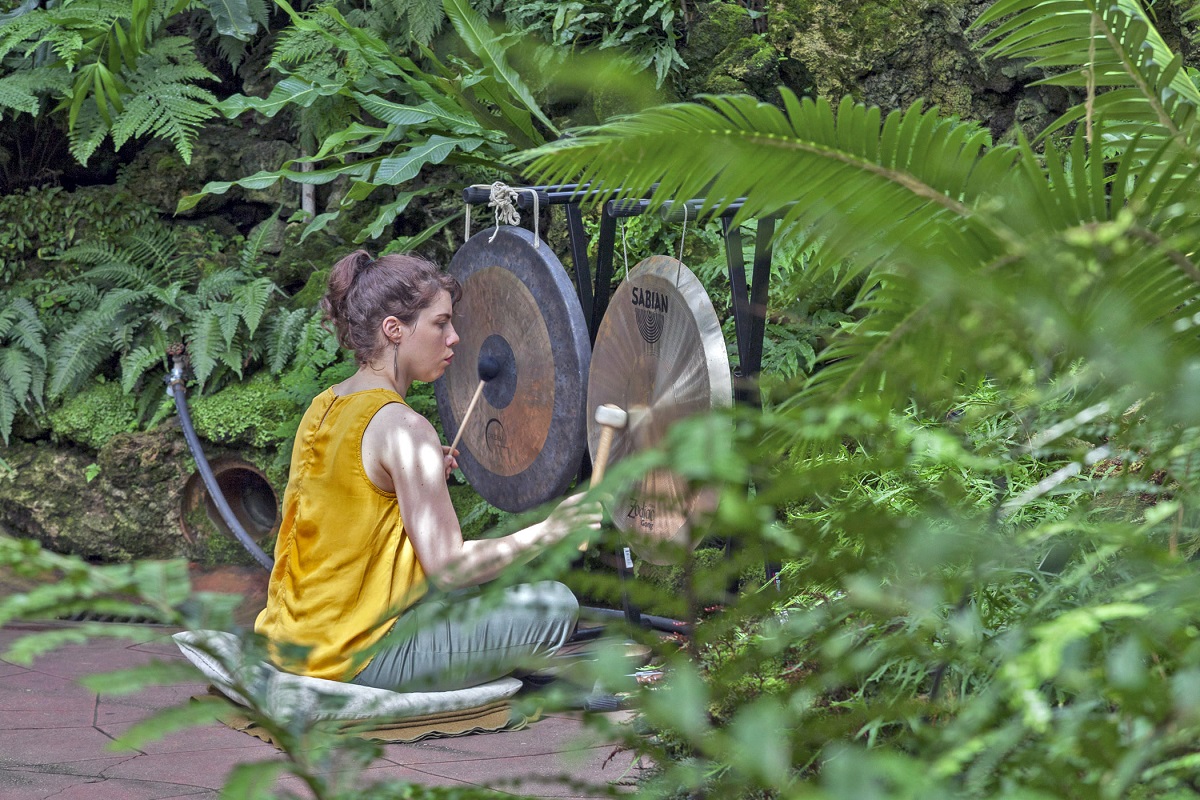 Woman in yellow top sits on cushion and hits two hanging gongs with mallets on a stone patio surrounded by foliage