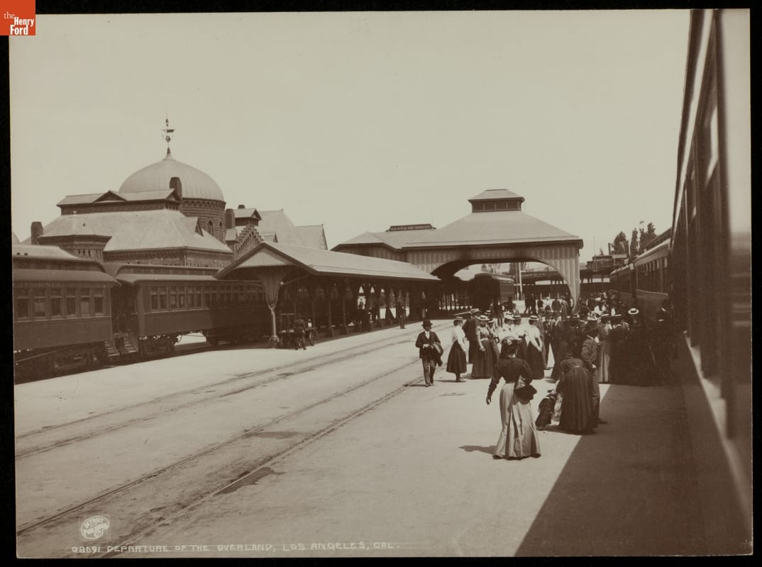 Crowd of people stands by railway tracks with trains on them and a building on the other side of the tracks
