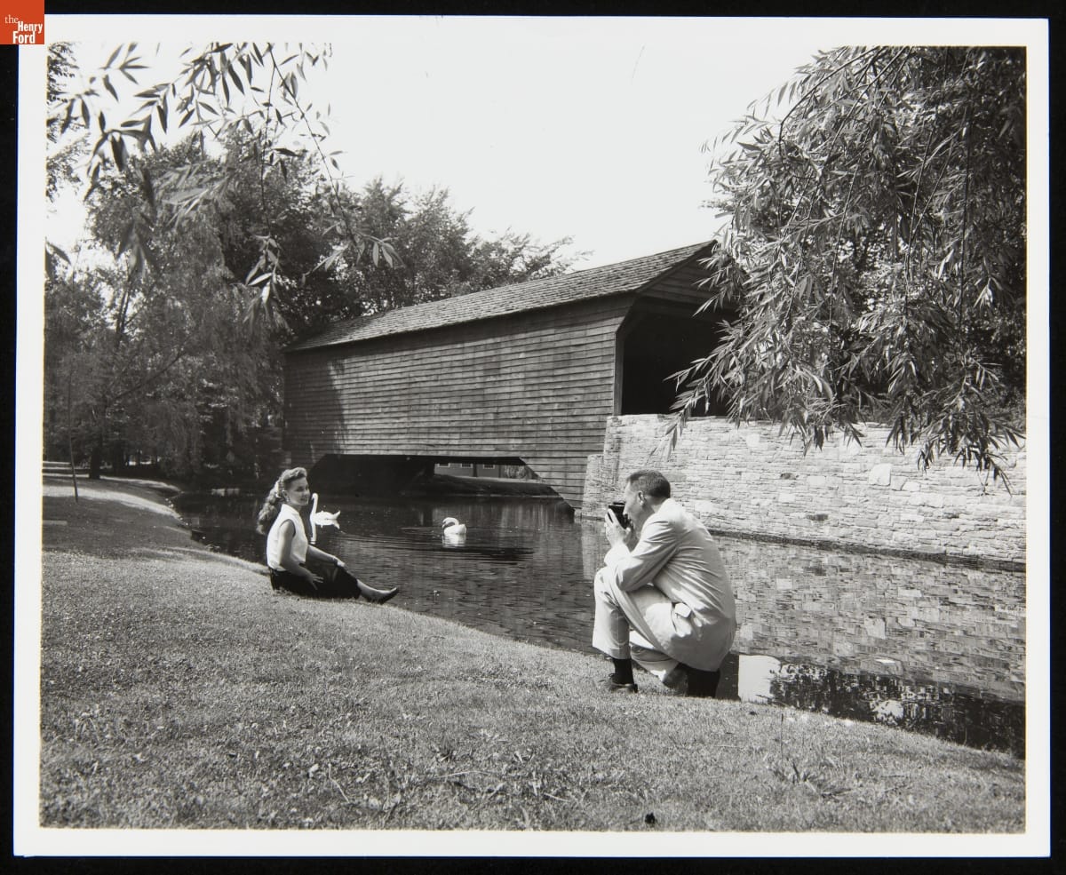 Lloyd Van Meter Photographing Nancy Lawrence near Ackley Covered Bridge in Greenfield Village, June 1958 A man, kneeling, photographs a woman sitting on the bank of a pond with a covered bridge in the background