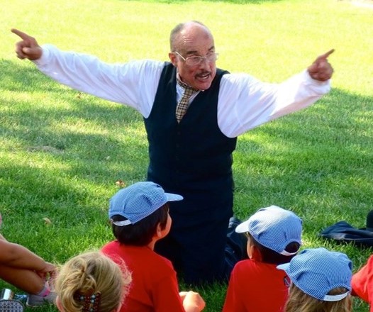 Man in suit (vest, pants, and white shirt) kneels on the ground with hands outstretched while children in baseball caps watch