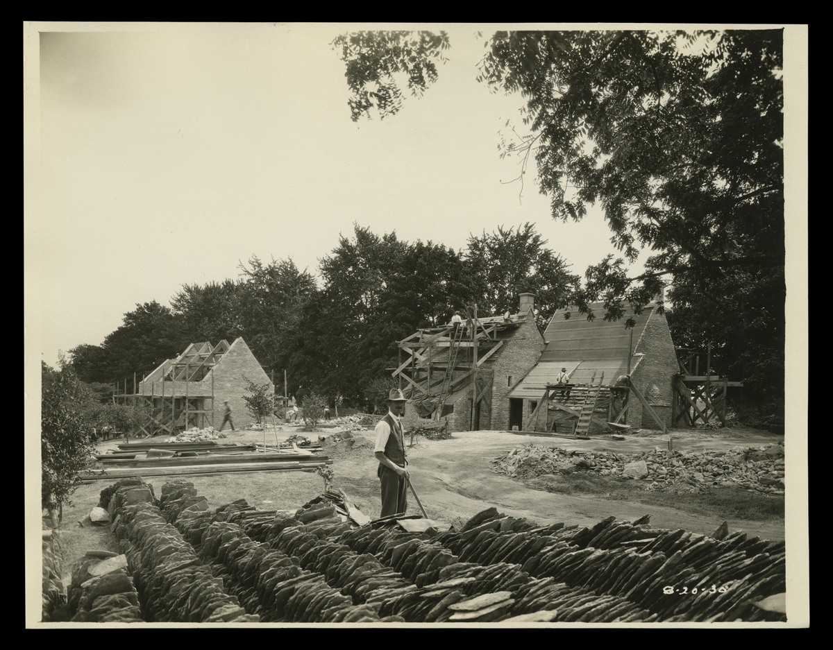 Man stands next to rows of stacked shingles in foreground; two partially constructed buildings in background