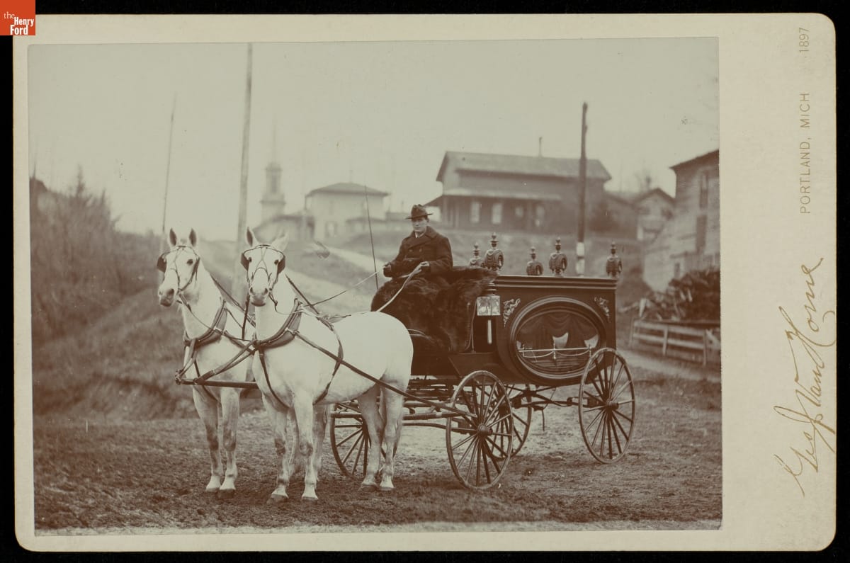 Horse-Drawn Hearse and Driver, 1897 Low carriage with oval glass window and seated driver, drawn by two horses, stopped in front of a road and houses