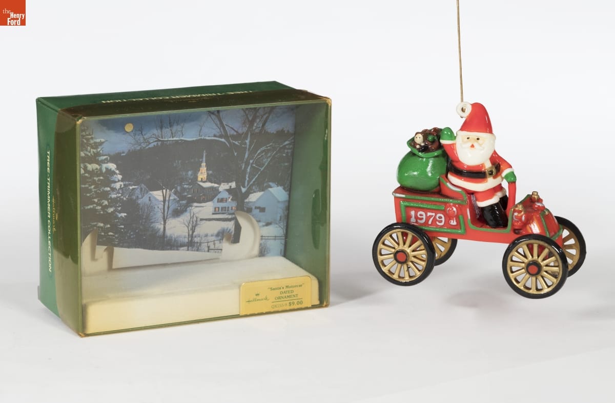 Ornament of Santa sitting in an old-fashioned open red car with "1979" on side and bag of toys in the backseat; a green box with clear plastic front for the ornament sits next to it