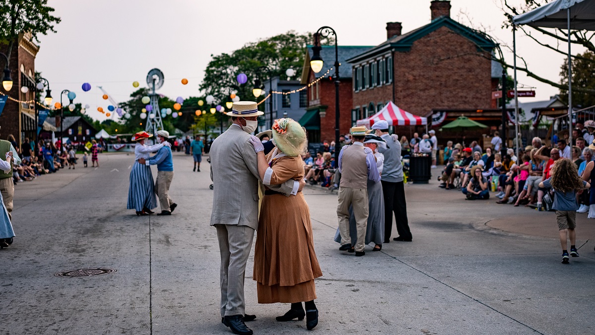 People wearing historical clothing dance in couples in a street as people look on from the sidelines