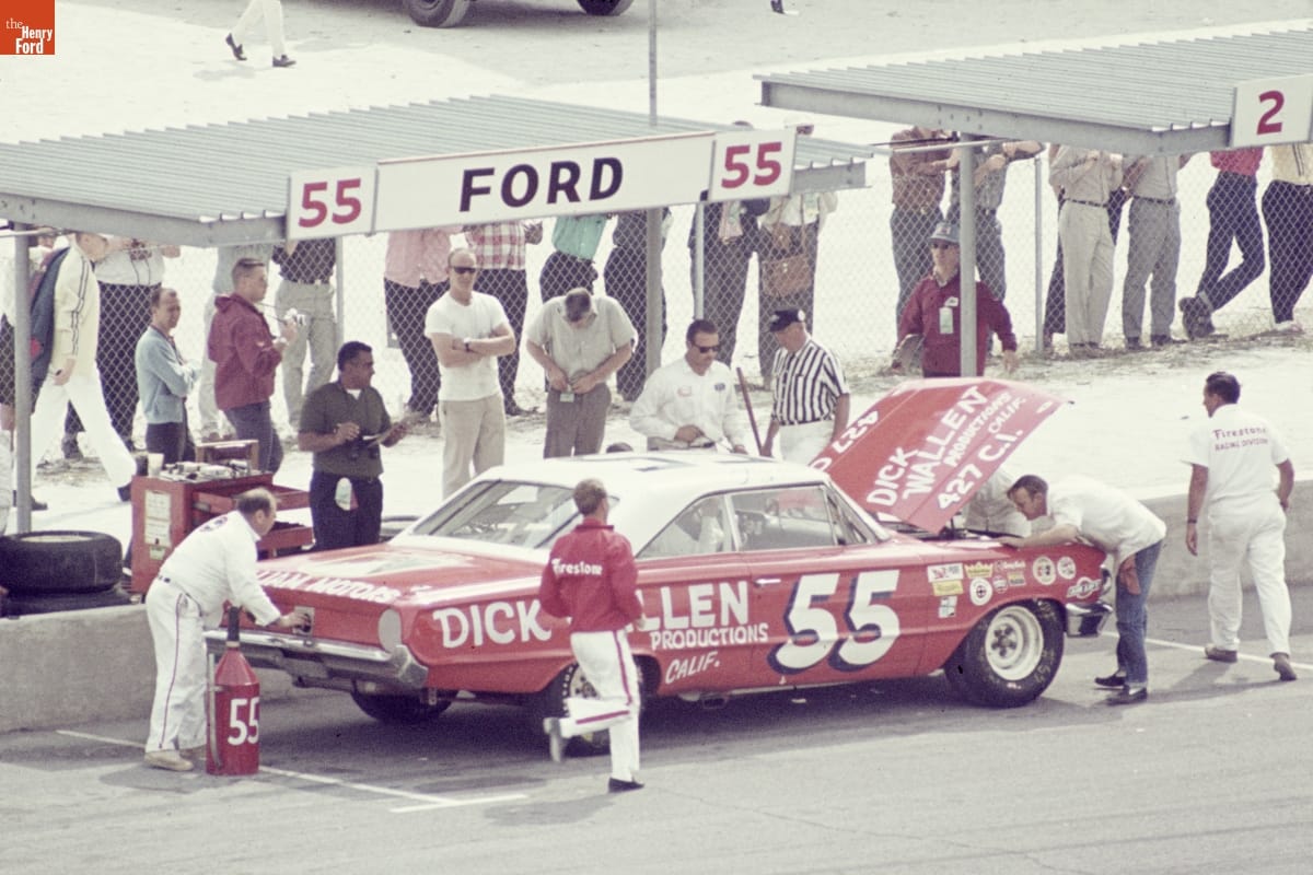 Daytona 500, February 14, 1965 Red car with text on sides, hood, and trunk is surrounded by several people, a couple working on the car