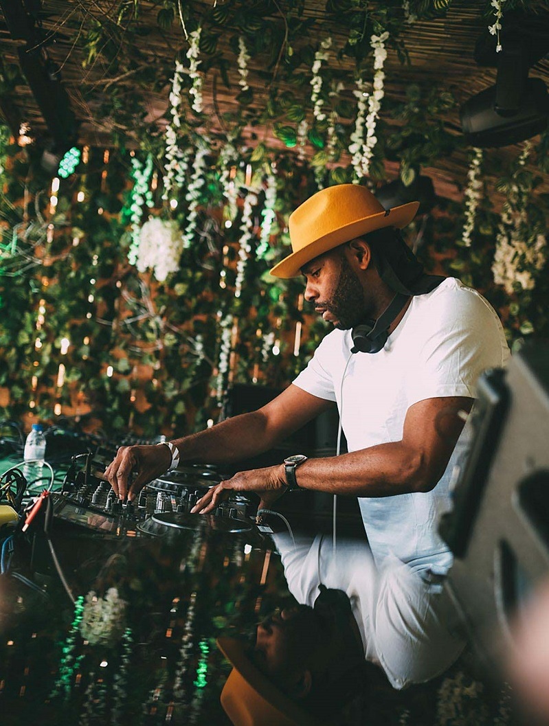 African American man in white t-shirt and yellow hat works at a DJ mixing station with foliage descending from wooden walls and ceiling in the background