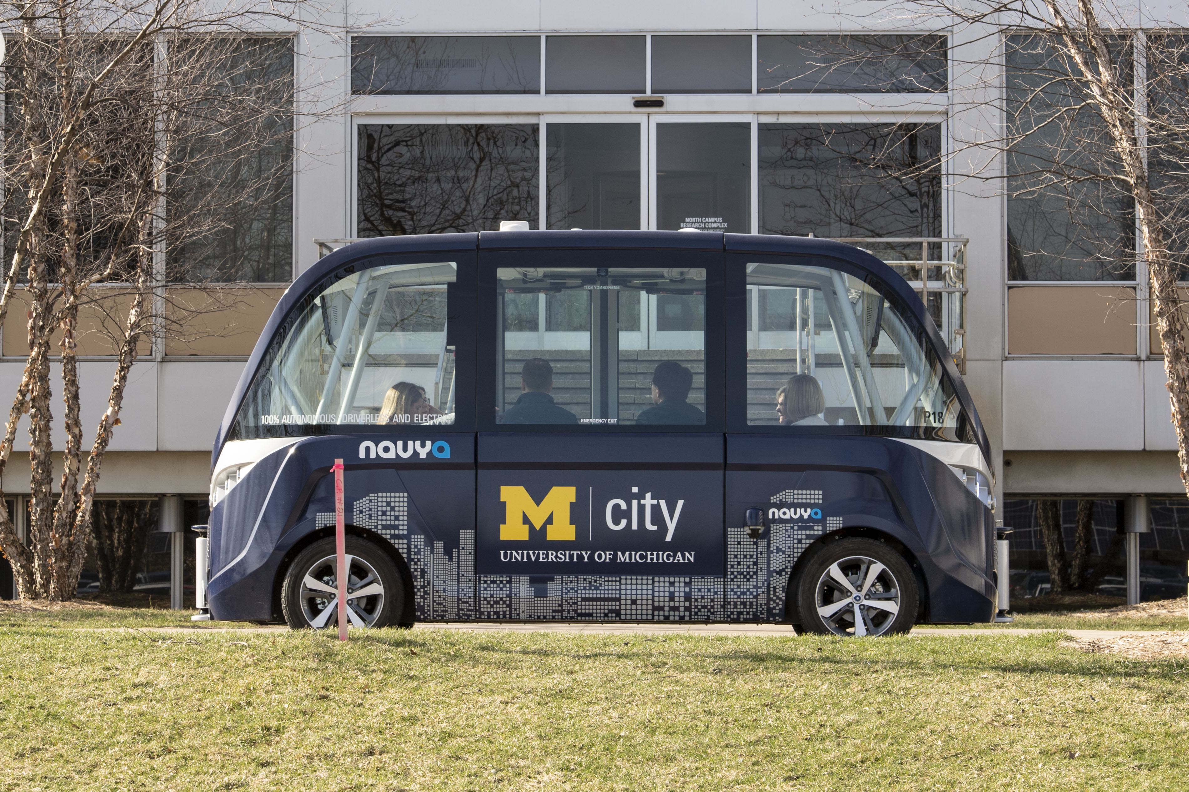 Side view of boxy blue shuttle with large windows and several people visible inside; grass in foreground and building in background