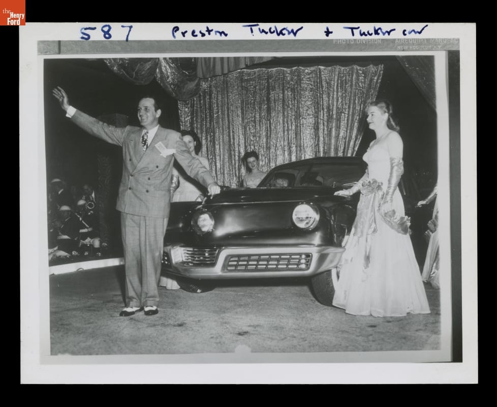 Black-and-white photo of man in suit and woman in fancy dress standing with two other women by a car in front of a curtain; man waves to crowd