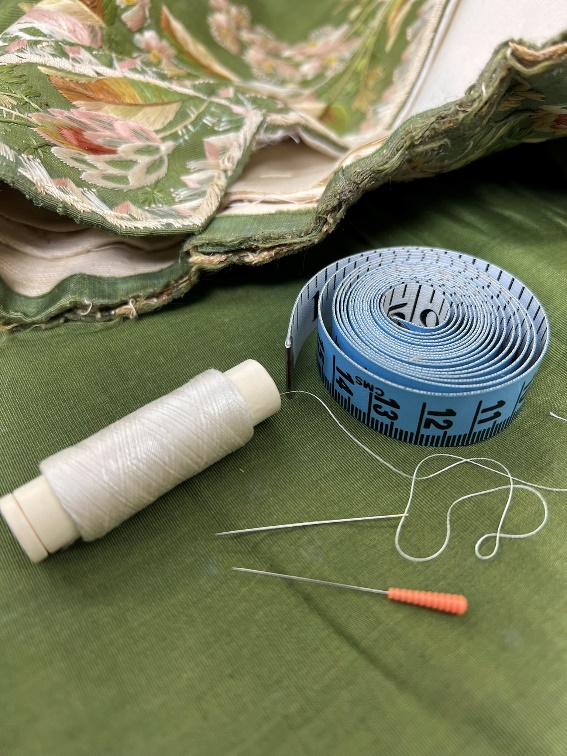 A tape measure, spool of white thread, needle, and small auger lay on top of moss-green fabric with elaborate embroidery