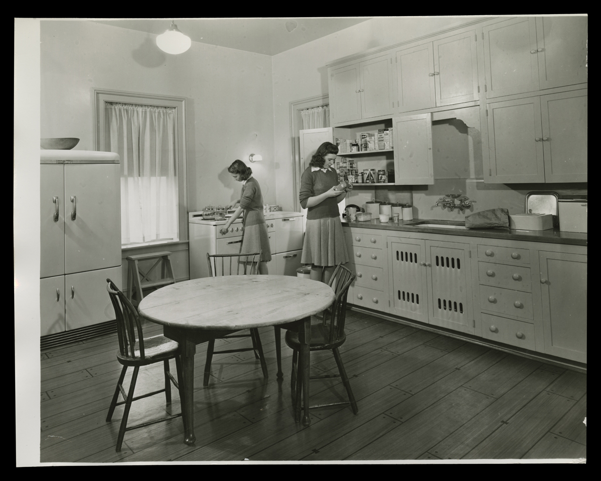 Edison Institute High School Students in Kitchen at Noah Webster Home, 1942 Two young women in a kitchen, one looking at a package from a cabinet and the other at a stove