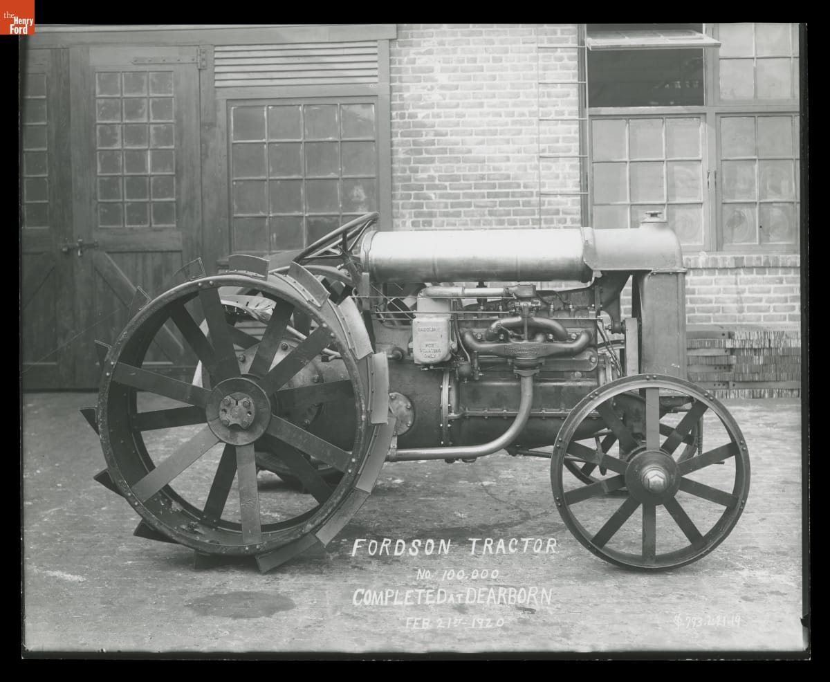 Black-and-white photo of a tractor sitting in front of a brick building with wooden doors and windows; also contains text
