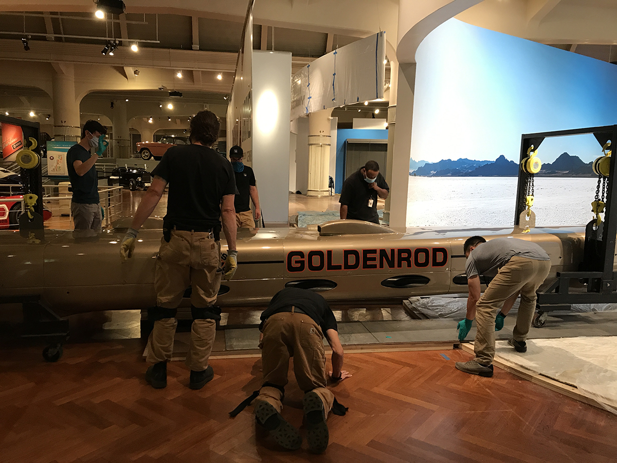 Group of men wearing facemasks stand and kneel around a long, low, torpedo-shaped golden car