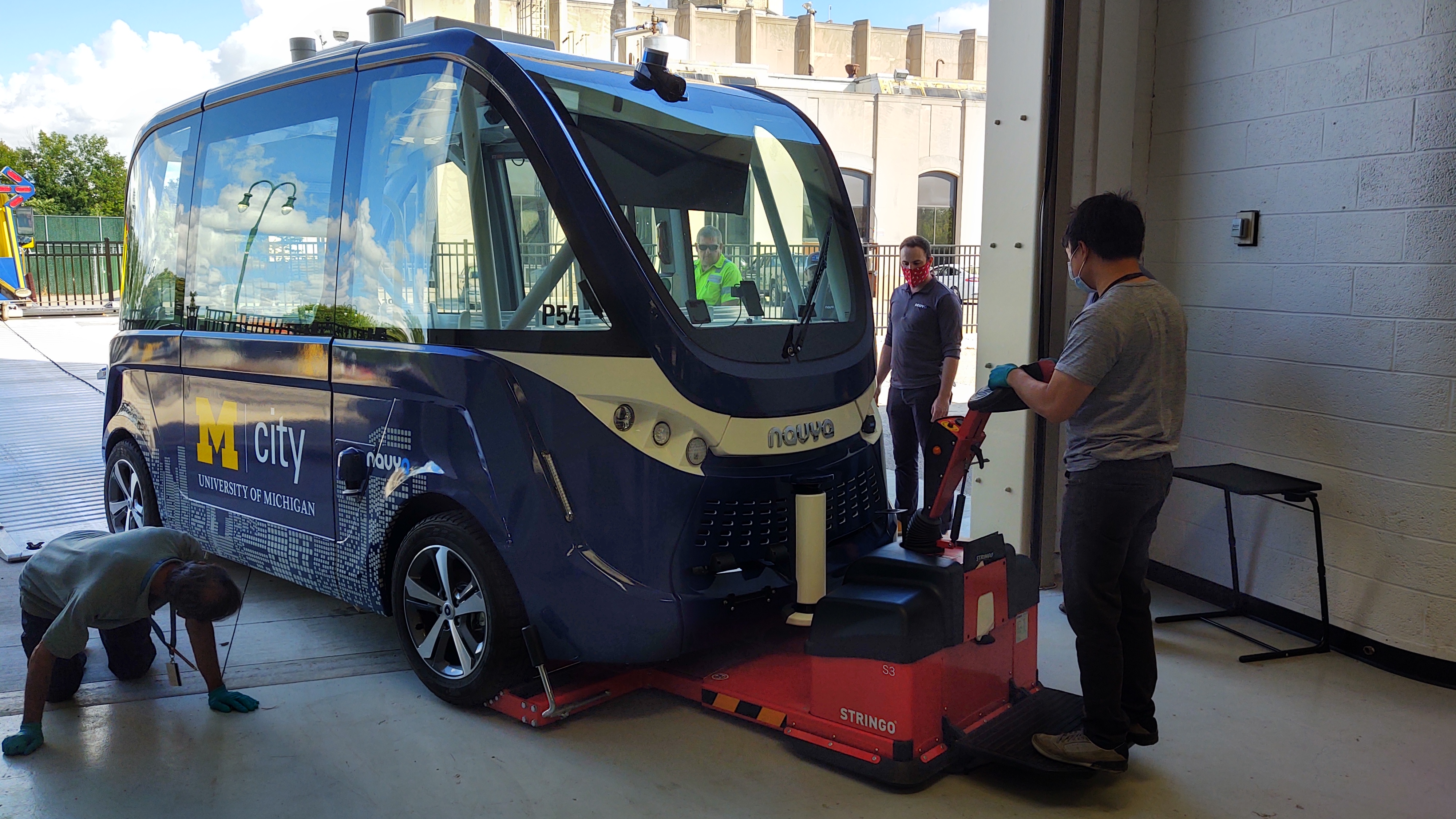 Mcity Driverless Shuttle arrives at The Henry Ford Boxy blue vehicle in doorway; one person on hands and knees looking underneath; 2 other people standing nearby