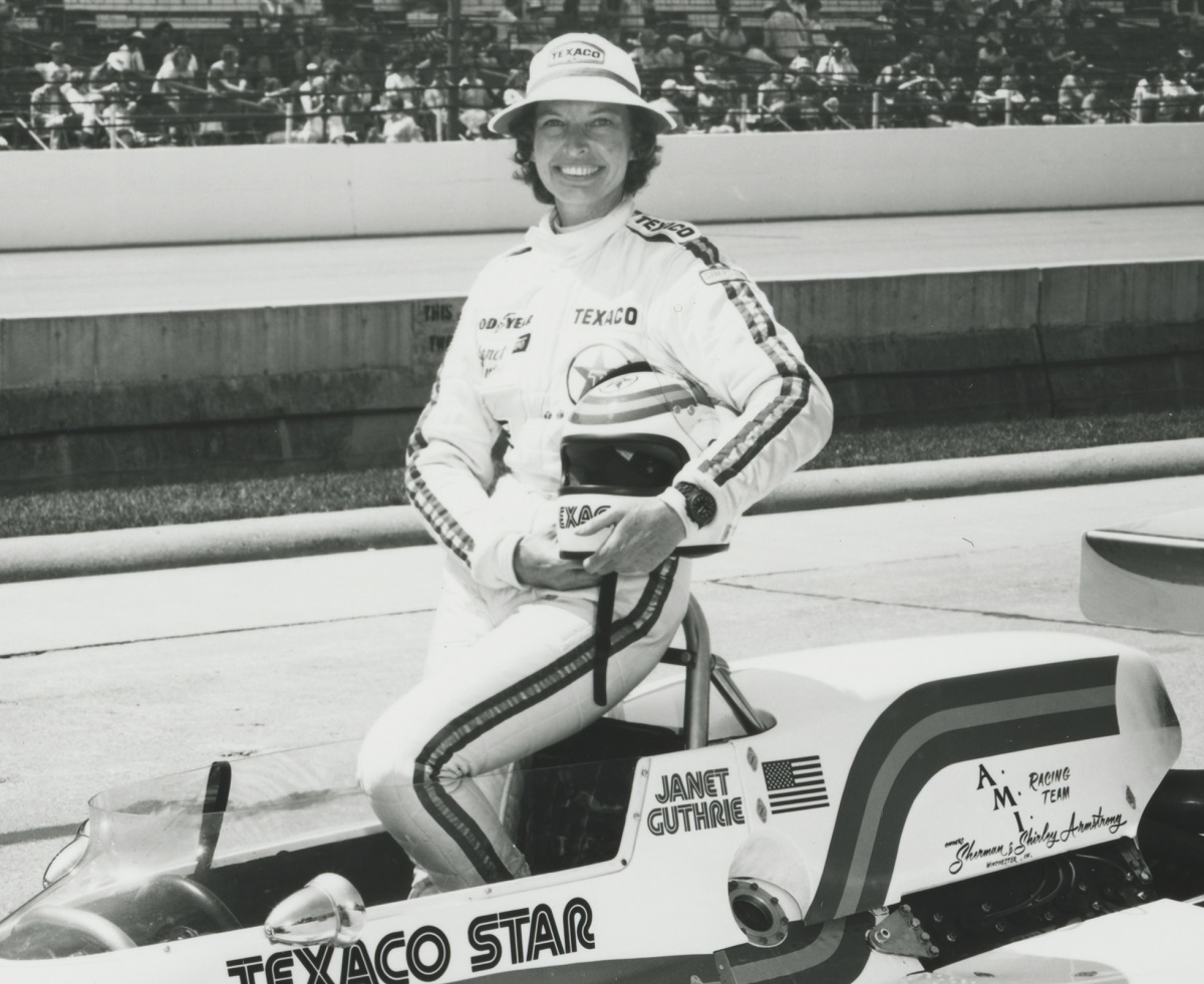 Janet Guthrie with Lola/Cosworth Race Car at Indianapolis Motor Speedway, 1979 (detail) Woman in racing jumpsuit holding helmet stands in race car on track with stands of people behind her