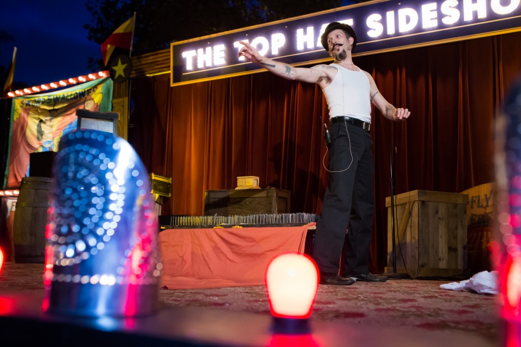 Man in black pants and hat and white tank top points from a stage, with lighted "Top Hat Side Show" sign behind him
