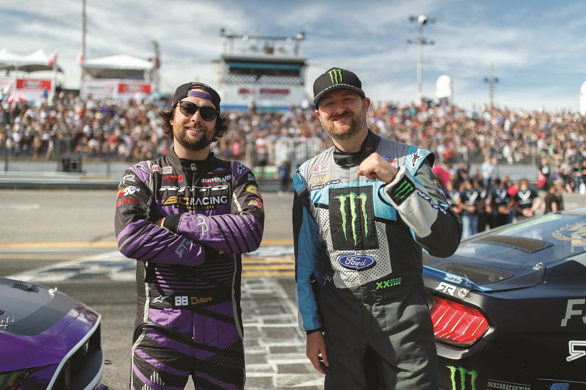 Two men in racing jumpsuits and baseball caps stand by cars in front of a track and spectators in grandstands