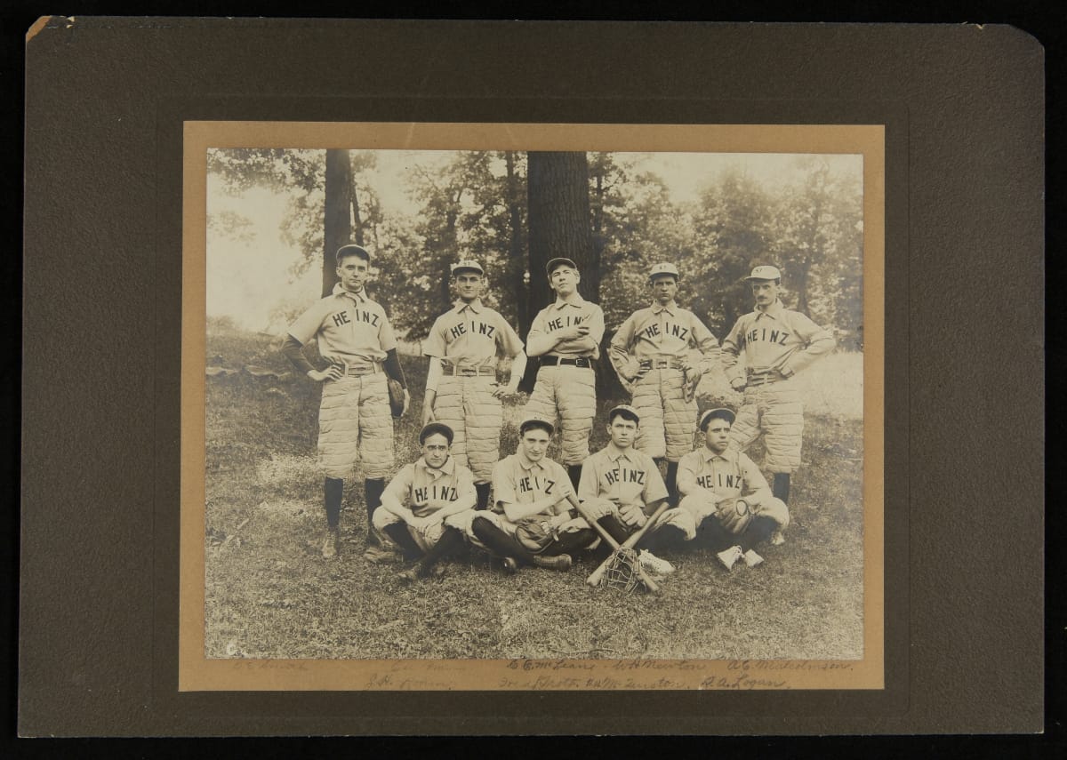 Black-and-white photo of five men standing, four men sitting, some with gloves and bats, wearing baseball uniforms and caps